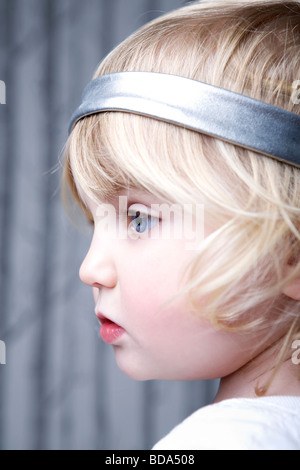 Un close up ritratto di una bambina con luminosi occhi blu. Lei ha i capelli biondi e indossa un cerchietto per capelli in argento e una camicia bianca. Foto Stock