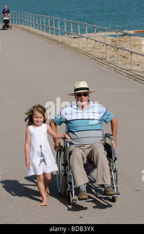 Bambina cammina con il suo nonno un uomo anziano in una sedia a rotelle sul lungomare Foto Stock