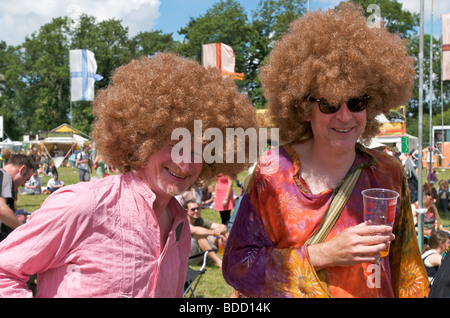 Camicie hippy e afro parrucche indossata da due uomini come fancy dress al Womad Festival di musica di gustare una birra sotto il sole Foto Stock