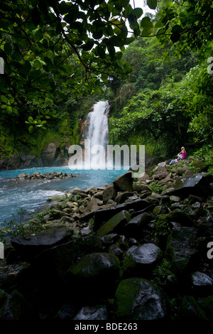 Escursionista femmina appoggiata al di sotto di una cascata lungo la vibrante blu Rio Celeste nel fiume Tenorio Il Parco Nazionale del Vulcano, Costa Rica. Foto Stock