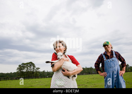Senior donna azienda baby capra in campo, marito in background Foto Stock
