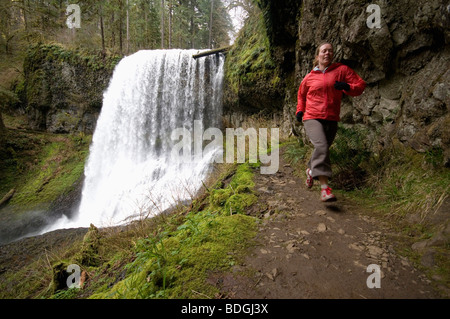 Una donna trail running accanto ad una cascata in Silver Falls State Park, Oregon, Stati Uniti d'America. Foto Stock