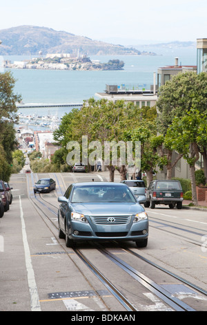 Vetture guida su una ripida strada inclinata nel centro di San Francisco (Alcatraz visibile in background) Foto Stock