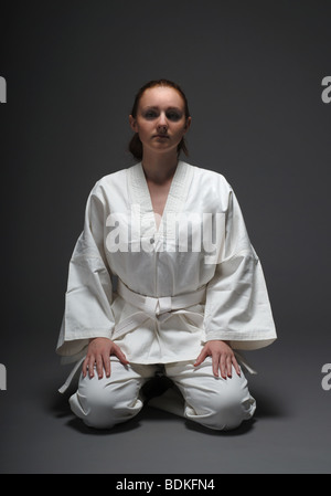 Ragazza in bianco uniforme si siede sulle gambe contro lo sfondo scuro, posa tradizionale Foto Stock