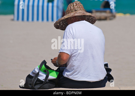Un uomo anziano che indossa un cappello di paglia per la protezione dal sole si siede su una parete in spiaggia guardando al suo Apple IPhone. Foto Stock