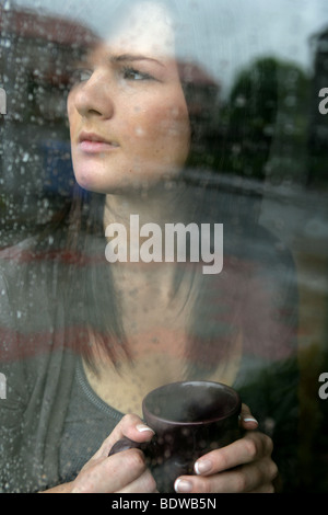 Dai capelli scuri donna tenendo una tazza di caffè guardando fuori una pioggia inzuppato finestra. Foto Stock