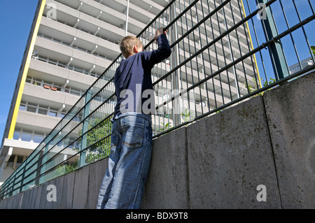 Il ragazzo, 9, guardando attraverso la recinzione metallica di un parco giochi privato nella parte anteriore di un alto edificio di appartamenti, città satellite di C Foto Stock