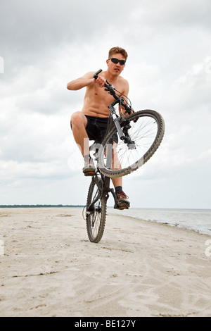 Giovane uomo facendo impennata con la bicicletta in spiaggia Foto Stock