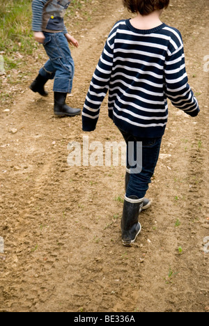 Vista ritagliata di 2 giovani ragazzi gambe in stivali da pioggia a piedi lungo una pista melmosa Foto Stock