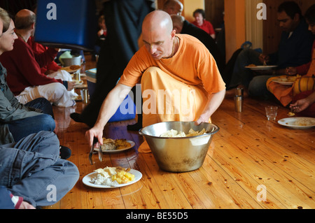 Hare Krishna discepolo, vestito in arancione abiti dello zafferano, serve cibo a una festa vegetariana in una stanza del tempio Foto Stock