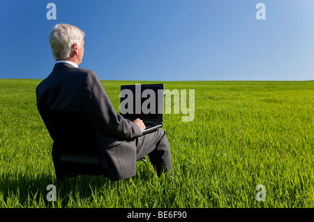 Il concetto di Business shot che mostra un vecchio Esecutivo maschio utilizzando un computer portatile in un campo verde con un cielo blu. Foto Stock
