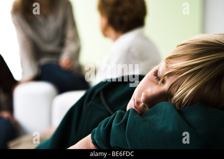 Ragazzo appoggiato la testa sulle braccia del sonno, membri della famiglia in background Foto Stock