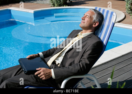 Imprenditore maturo in appoggio sulla sedia a sdraio accanto alla piscina tenendo il computer portatile Foto Stock