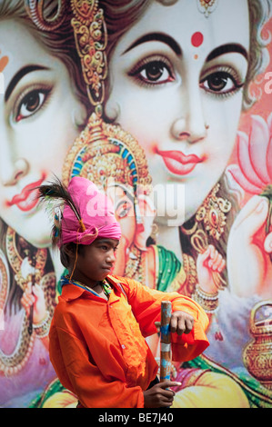 Mendicante indiano ragazzo con lo strumento musicale contro un poster di divinità Indù. Andhra Pradesh, India Foto Stock