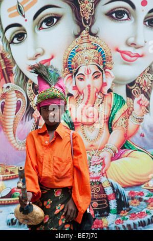 Mendicante indiano ragazzo con lo strumento musicale contro un poster di divinità Indù. Andhra Pradesh, India Foto Stock