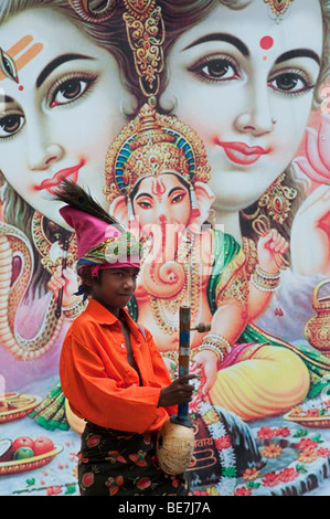 Mendicante indiano ragazzo con lo strumento musicale contro un poster di divinità Indù. Andhra Pradesh, India Foto Stock