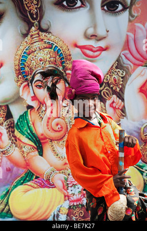 Mendicante indiano ragazzo con lo strumento musicale contro un poster di divinità Indù. Andhra Pradesh, India Foto Stock