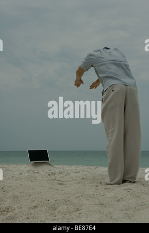 Uomo in piedi sulla spiaggia, piegarsi verso il computer portatile, vista posteriore Foto Stock