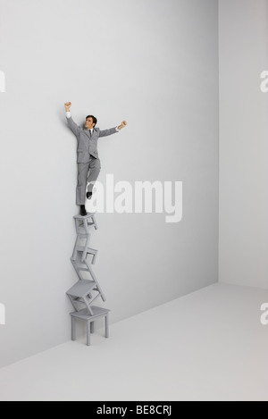 Imprenditore in piedi con le braccia alzate in cima a pila di sedie Foto Stock