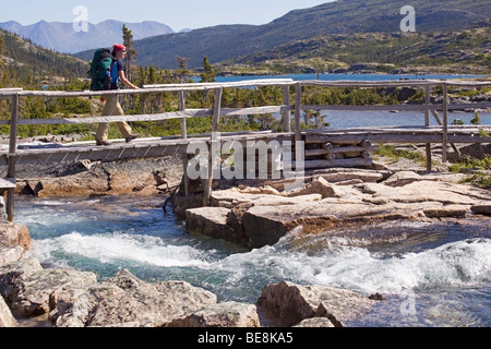 Giovane donna attraversando creek, ponte di legno, Lago Profondo dietro, escursionismo, backpacking, escursionista con zaino, storico Chilkoot Trail, Foto Stock