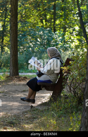 Donna anziana siede sulla panchina del parco e legge un libro religioso Foto Stock