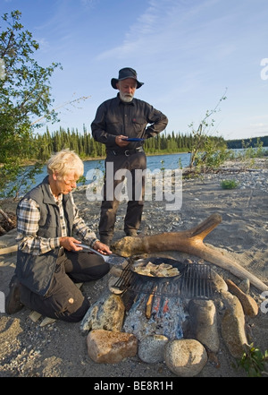 Donna di cottura, friggere i filetti di pesce in una padella su un fuoco di campo, l'uomo con la piastra in attesa dietro, superiore Liard River, Yukon Territory, Foto Stock