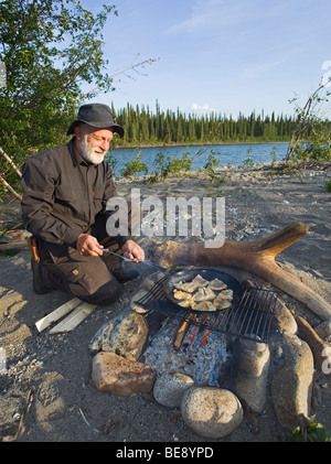 Uomo di cottura, friggere i filetti di pesce in una padella su un fuoco di campo superiore del Fiume Liard, Yukon Territory, Canada Foto Stock