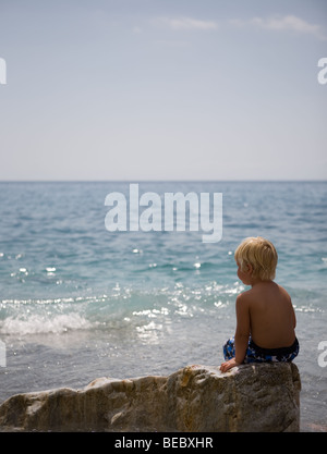 Ragazzino che guarda il bellissimo mare mentre è seduto su una roccia Foto Stock