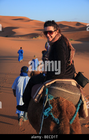 Turista femminile su un cammello trek guidato da uomo blu attraversa Erg Chebbi dunesin di sabbia del deserto del Sahara vicino a Merzouga, Marocco Foto Stock