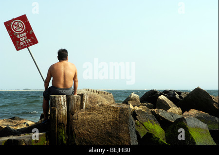 Un uomo si siede su un pontile che guarda all'oceano sulla spiaggia a Coney Island, Brooklyn, New York, Stati Uniti d'America. Foto Stock