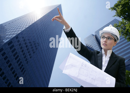 Bella giovane architetto ingegnere con hardhat e piani di fronte urbano della città sullo sfondo di edifici Foto Stock