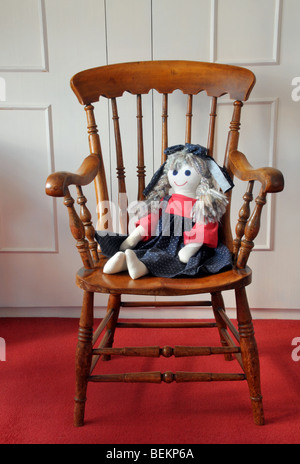 Un fatto in casa di bambola di stoffa Vestito in blu e rosso pinafore con un inchino nei suoi capelli, si siede su una sedia in legno. Foto Stock