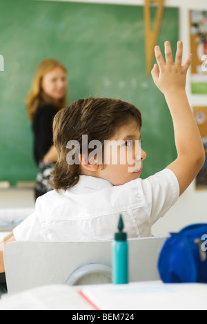 Ragazzo alzando la mano in classe, guardando sopra la spalla Foto Stock
