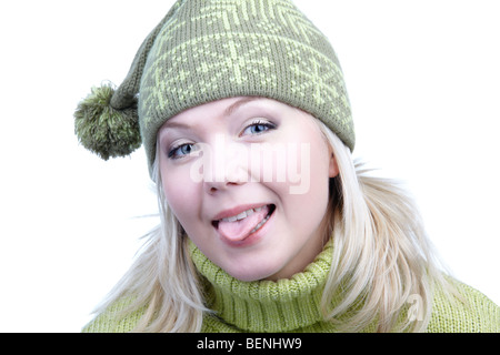 Ritratto di giocoso bella ragazza bionda in maglione verde e cappello invernale con bob mettendo fuori la sua lingua Foto Stock