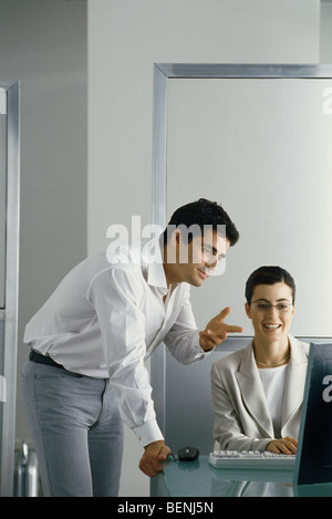 Office scena, uomo appoggiata sul bordo della scrivania che parla al collega di sesso femminile, sia guardando il suo schermo del computer Foto Stock