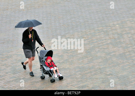 Papà con ombrellone e baby nella PRAM camminare sul lungomare lungo la costa in località balneare sul giorno di pioggia durante le vacanze estive Foto Stock