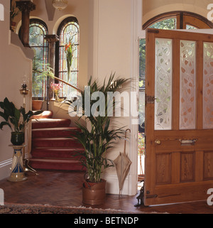 Houseplants verde su entrambi i lati della scala in crema hall con porta anteriore aperta con inciso i pannelli in vetro Foto Stock