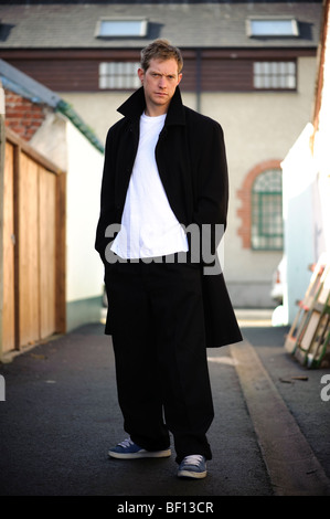 30 anni di vecchio uomo in piedi da solo in un vicolo con le mani in tasca che indossa mantello nero t-shirt bianco sneakers, Regno Unito Foto Stock