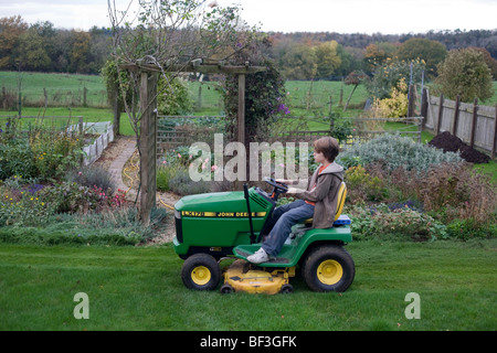 Un undici anno-vecchio ragazzo aziona suo nonno tosaerba trattore da solo nel giardino della famiglia. Foto Stock