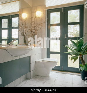 Luce da parete a parete accanto a doppia vetrata porte in moderno bagno in piastrelle bianche con montato lavabo e wc su piastrelle unità dello specchietto di cortesia Foto Stock