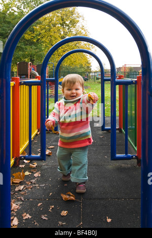 Ritratto verticale di una bambina toddling attraverso un elemento tubolare a labirinto con foglie cadute in mano a una zona giochi per bambini. Foto Stock