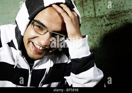 Ritratto di un giovane uomo sorridente mentre indossa una giacca con cappuccio seduto di fronte a un vecchio sportello in acciaio, gioventù, guardando direttamente a Foto Stock