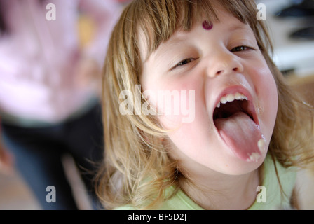 Giovane ragazza di tre anni, con torte sulla sua lingua dopo il leccare il cucchiaio di miscelazione. Foto Stock