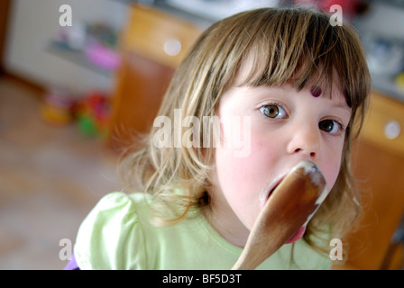 Ragazza giovane, tre anni, leccare il cucchiaio dopo aver aiutato per mescolare una torta. Foto Stock