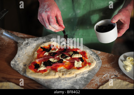 Aggiunta di ingredienti a casa una pizza fatta in una cucina Foto Stock