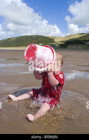 Un bambino in una muta che versa un secchio d'acqua sopra se stesso sulla spiaggia in estate Foto Stock