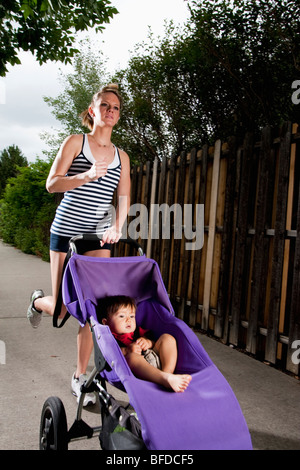 Un tagliente-cercando, Atletica Giovane donna gode di una corsa su un marciapiede suburbana con un bambino in un passeggino per fare jogging. Foto Stock