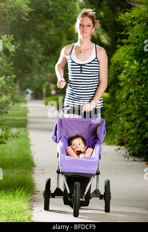 Un tagliente-cercando, Atletica Giovane donna gode di una corsa su un marciapiede suburbana con un bambino in un passeggino per fare jogging. Foto Stock