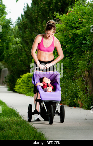 Un tagliente-cercando, Atletica Giovane donna guarda verso il basso in corrispondenza di un bambino sorridente nel suo jogging passeggino durante una corsa su un marciapiede suburbana. Foto Stock