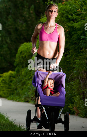 Baby solleva pugno sorridente in jogging passeggino spinto da un tagliente-cercando, Atletica Giovane donna durante una corsa su un marciapiede suburbana Foto Stock